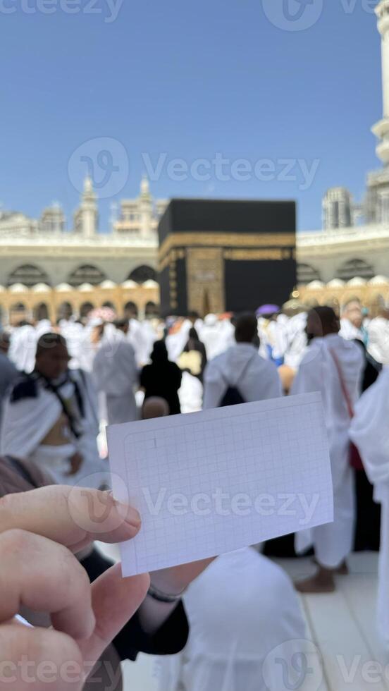 A pilgrim holds a small notebook with a heart doodle in front of the Kaaba in Mecca, Saudi Arabia. The sacred site is surrounded by worshippers performing Hajj or Umrah under a clear blue sky photo