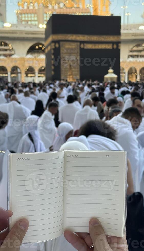 A pilgrim holds a small notebook with a heart doodle in front of the Kaaba in Mecca, Saudi Arabia. The sacred site is surrounded by worshippers performing Hajj or Umrah under a clear blue sky photo