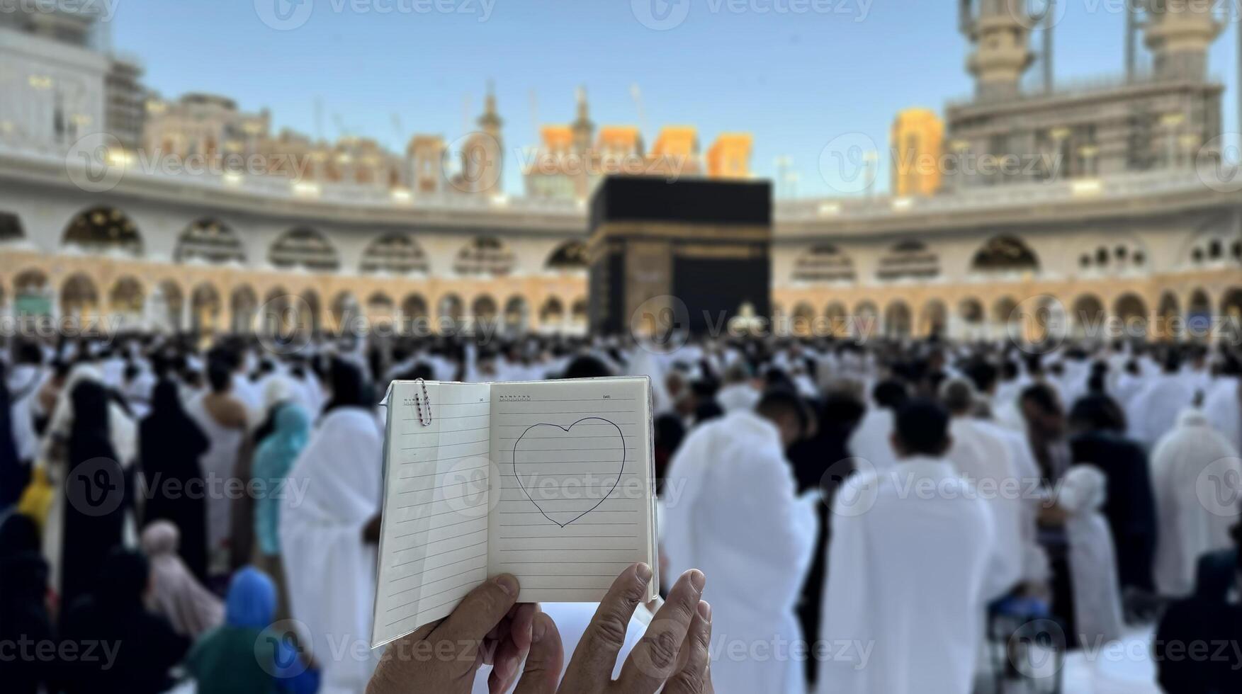 A pilgrim holds a small notebook with a heart doodle in front of the Kaaba in Mecca, Saudi Arabia. The sacred site is surrounded by worshippers performing Hajj or Umrah under a clear blue sky photo