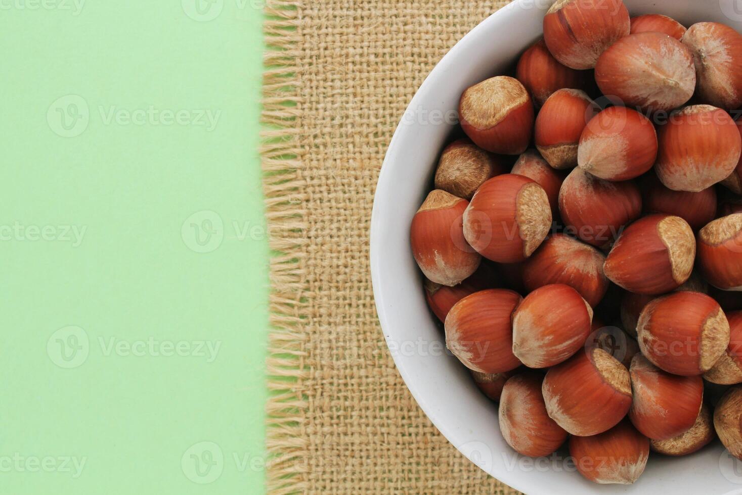 Hazelnuts in white bowl on green background. Bowl of hazelnuts with burlap border. Nuts in white bowl on green backdrop. Pile of hazelnuts in white container. Hazelnut in bowl with jute fabric photo