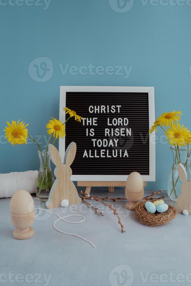 CHRIST THE LORD IS RISEN TODAY ALLELUIA text on letter board with bunny wooden figures eggs and fresh flowers. Easter composition content still life. Christian symbols of spring holiday photo