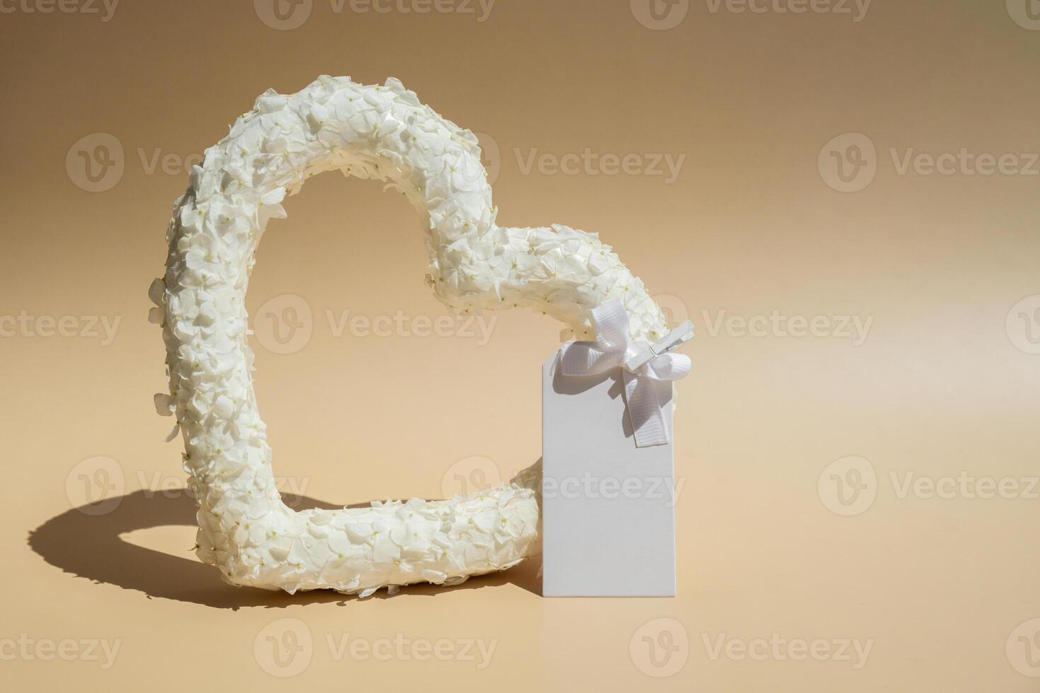Heart made of white lilac flowers with white mock up paper template on pastel beige background. Still life romantic composition of Valentines day holiday copy space for your text invitation greeting card postcard. photo