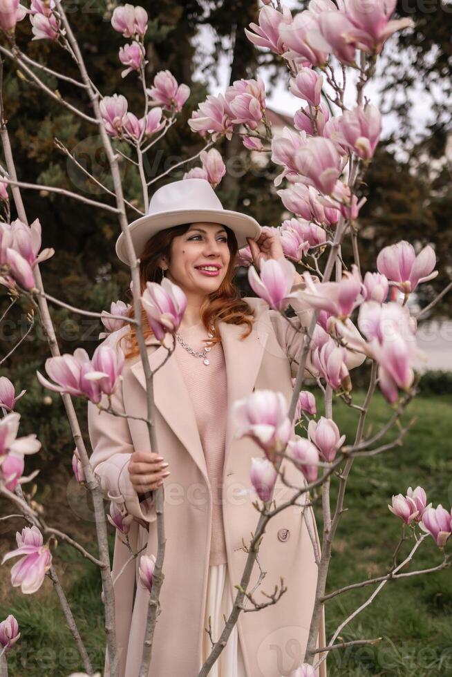 Woman magnolia flowers, surrounded by blossoming trees., hair down, white hat, wearing a light coat. Captured during spring, showcasing natural beauty and seasonal change. photo