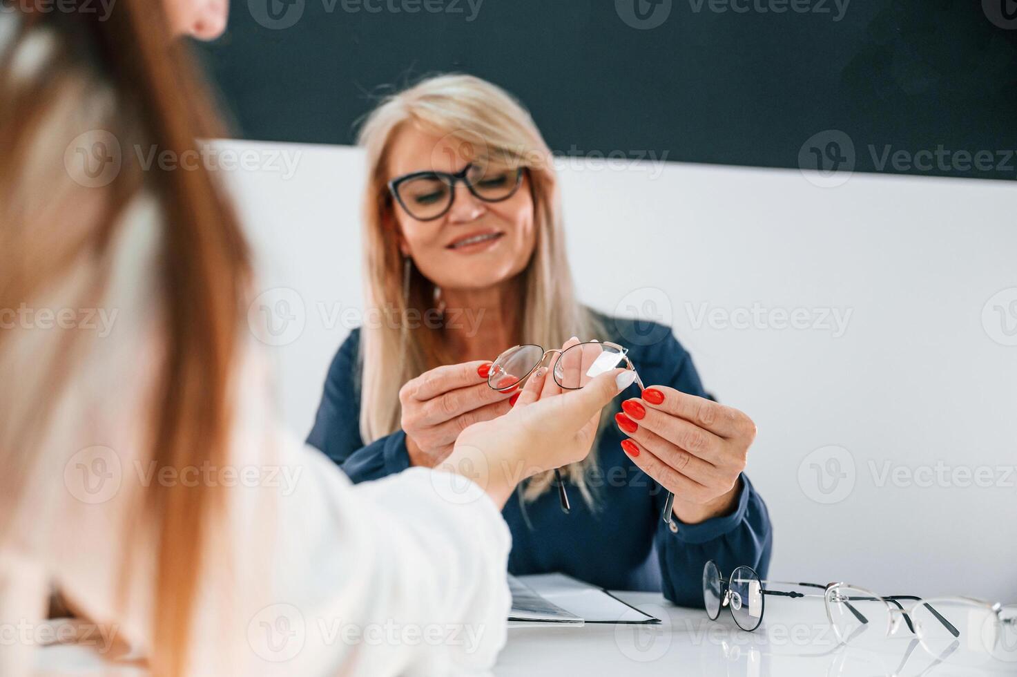 Sitting by the table. Woman choosing glasses in the store and getting help by assistant photo