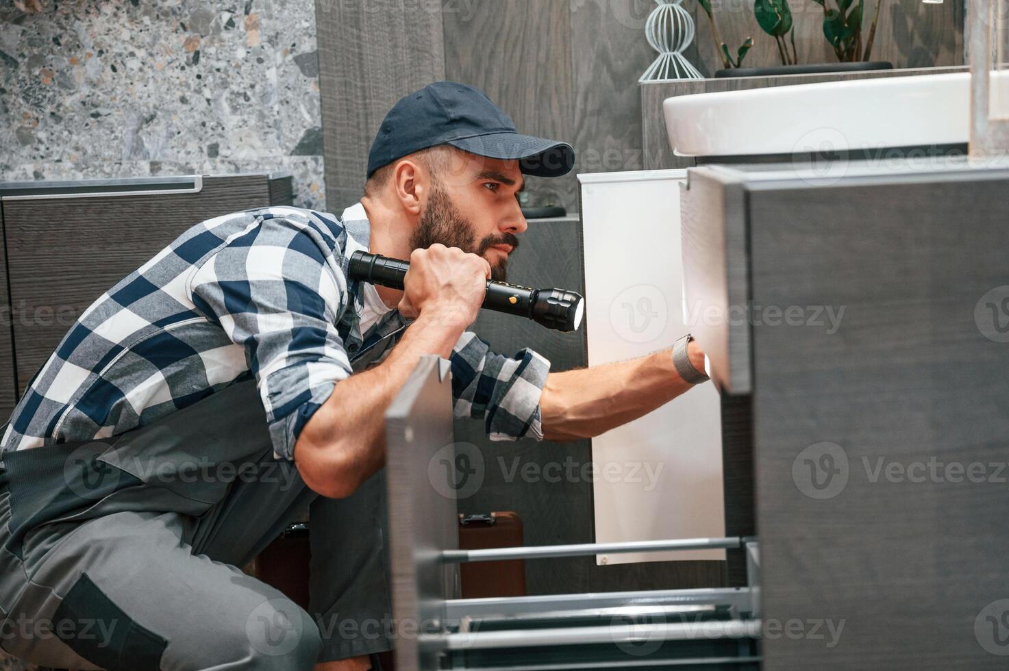 Using, flashlight. Plumber in uniform is indoors at home photo