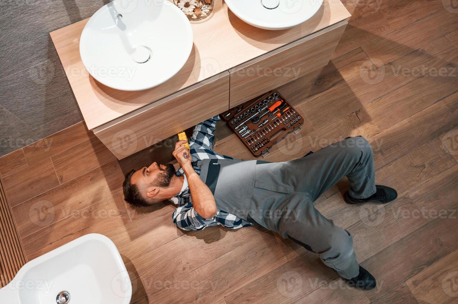 Laying down on the ground, fixing the sink. Plumber in uniform is indoors at home photo