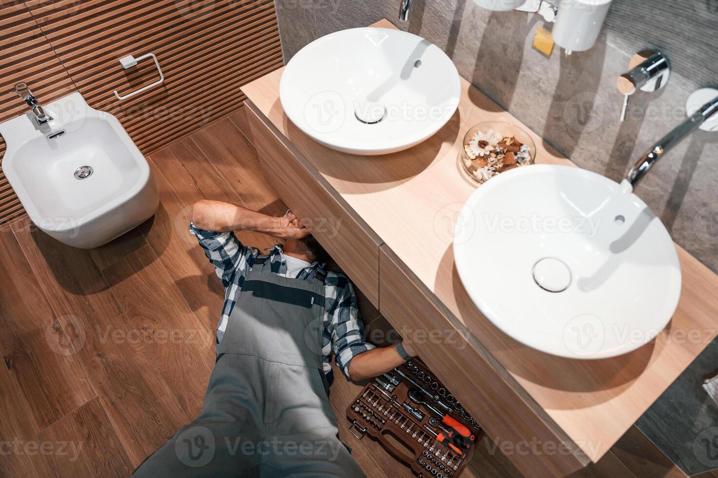 Laying down on the ground,, fixing the sink. Plumber in uniform is indoors at home photo