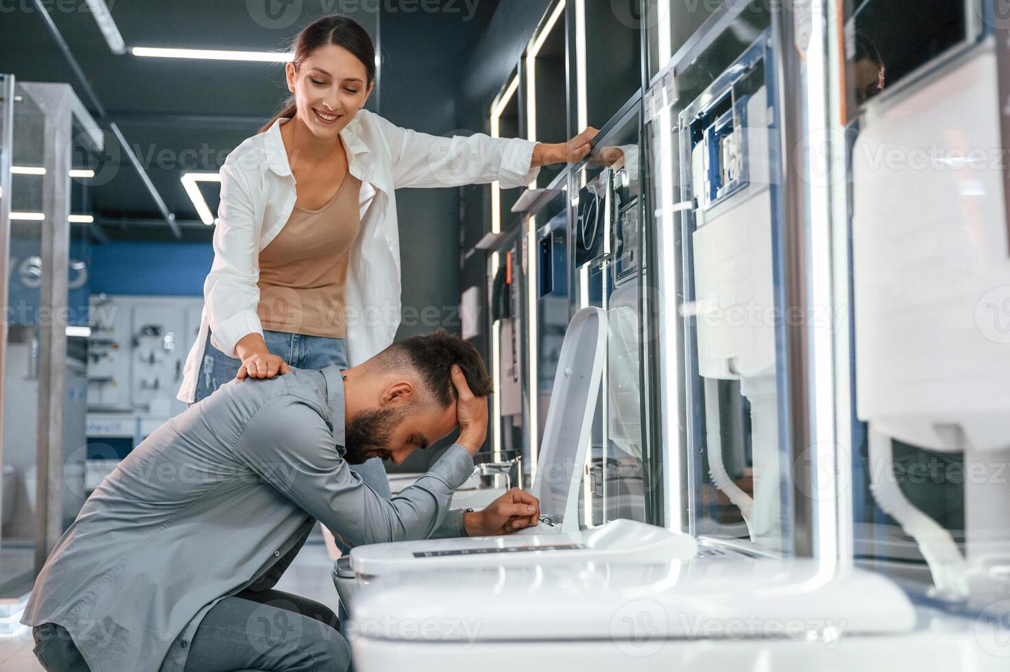 Man pretending he's, feeling bad at the toilet. Woman and man is in the store of goods for the home photo