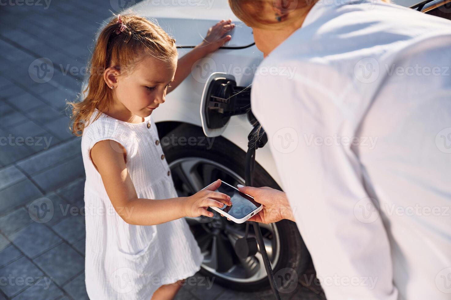 Using smartphone. Young woman with her little daughter is with their electric car outdoors photo