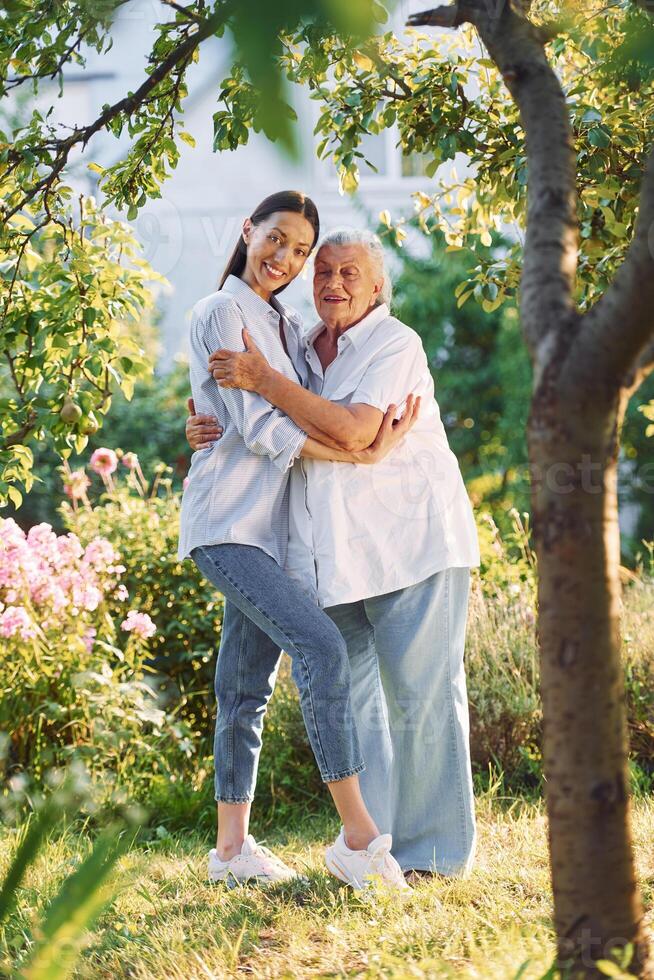 Embracing and posing, for a camera. Young woman is with her senior mother is in the garden photo