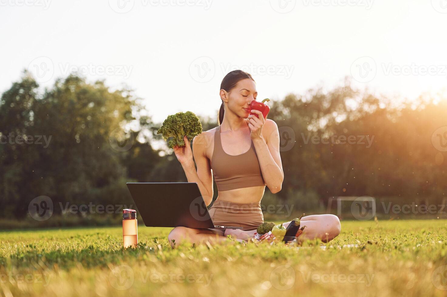 Using laptop.. Holding vegetables. Young woman in yoga clothes is outdoors on the field photo