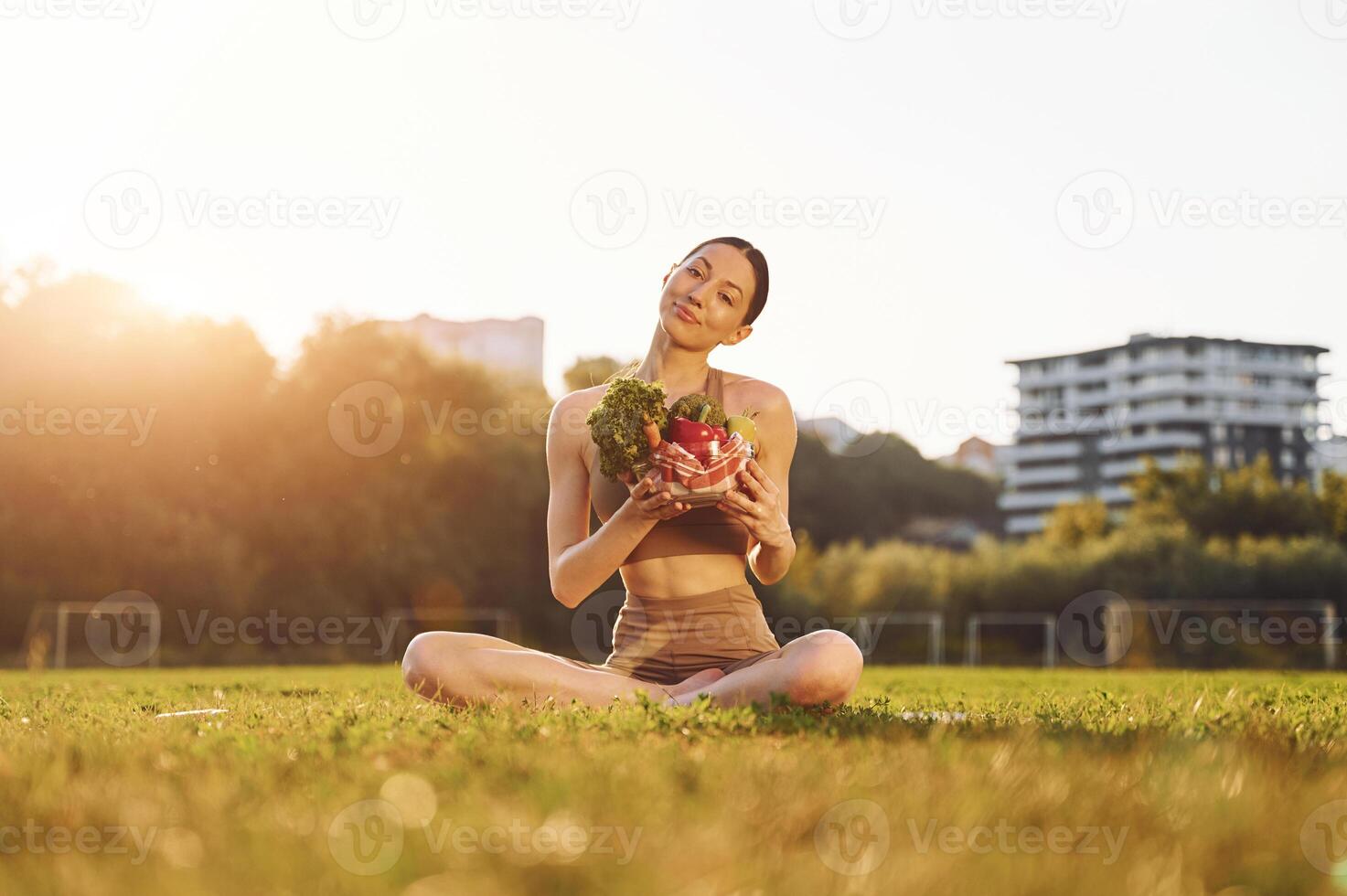 Sitting ,and holding plate with vegetables. Young woman in yoga clothes is outdoors on the field photo