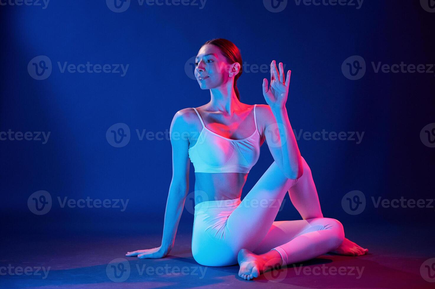 Sitting, on the floor and doing exercises. Young woman in sportive clothes is in the studio with neon lights photo