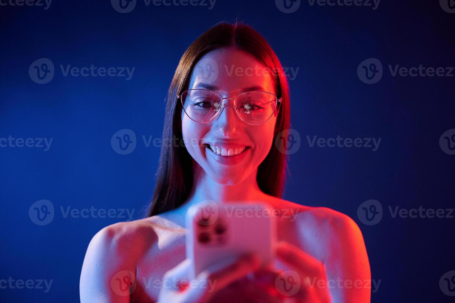 Holding, smartphone, and smiling. Portrait of young woman that is indoors in neon lighting photo