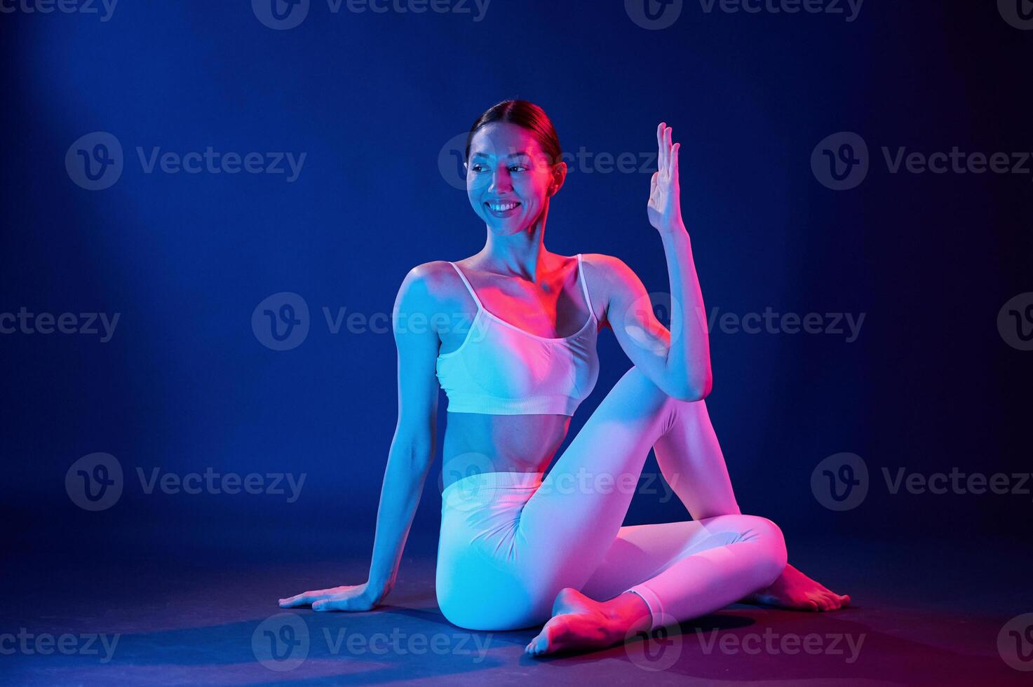 Sitting ,on the floor and doing exercises. Young woman in sportive clothes is in the studio with neon lights photo
