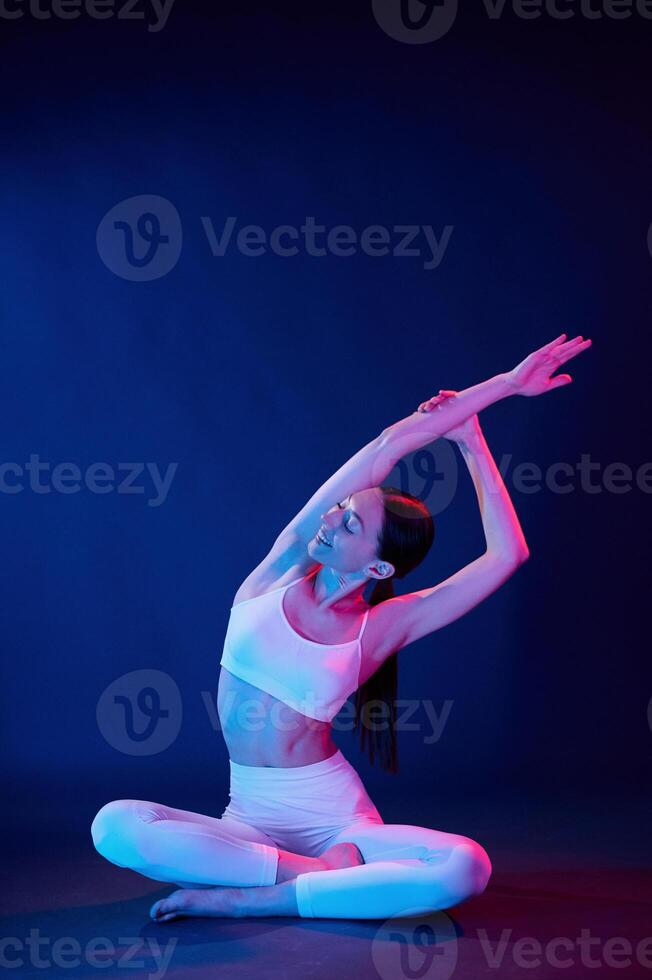 Doing exercises in lotus pose. Young woman in sportive clothes is in the studio with neon lights photo