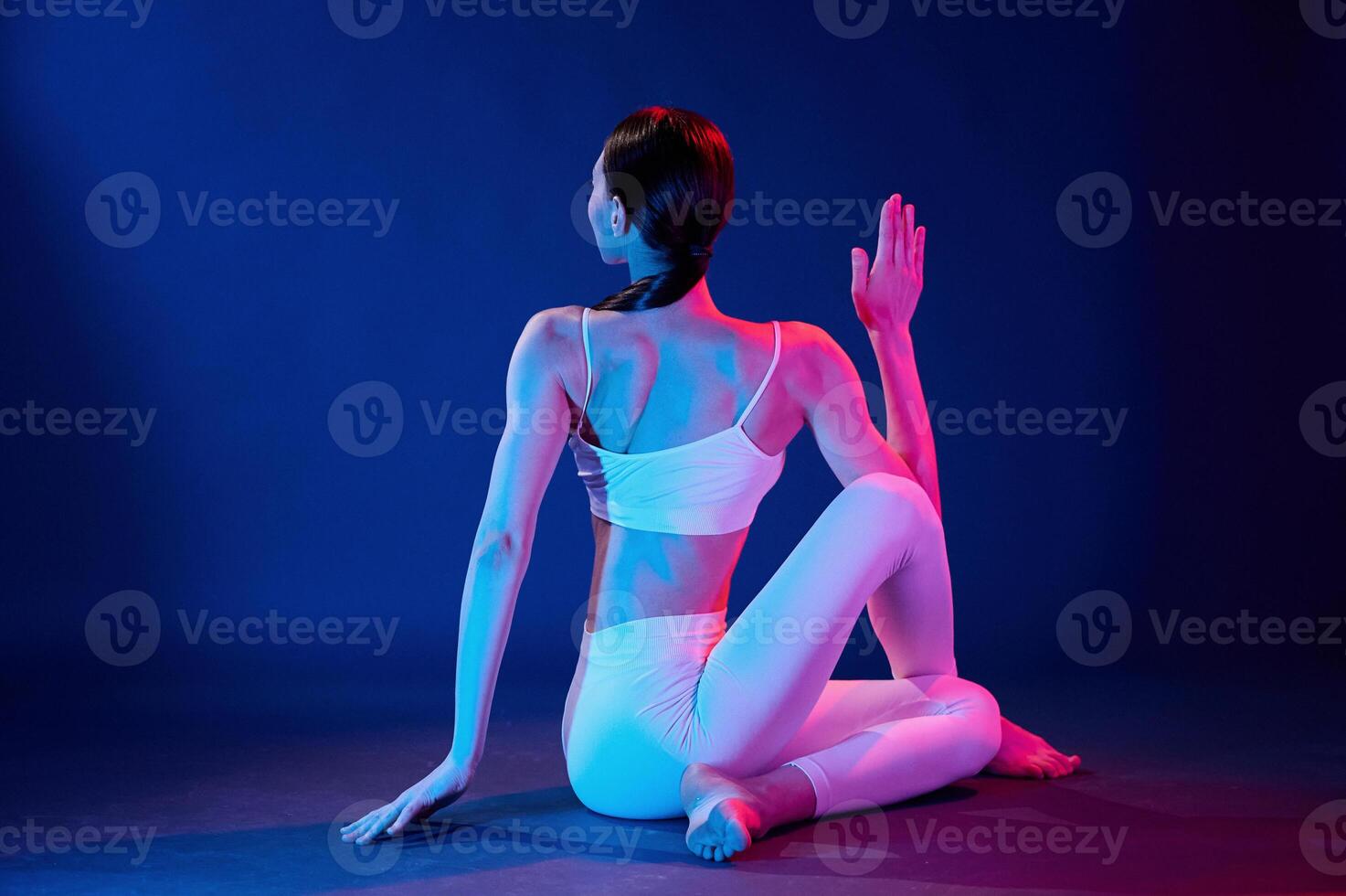 Rear view.. Sitting and doing exercises. Young woman in sportive clothes is in the studio with neon lights photo