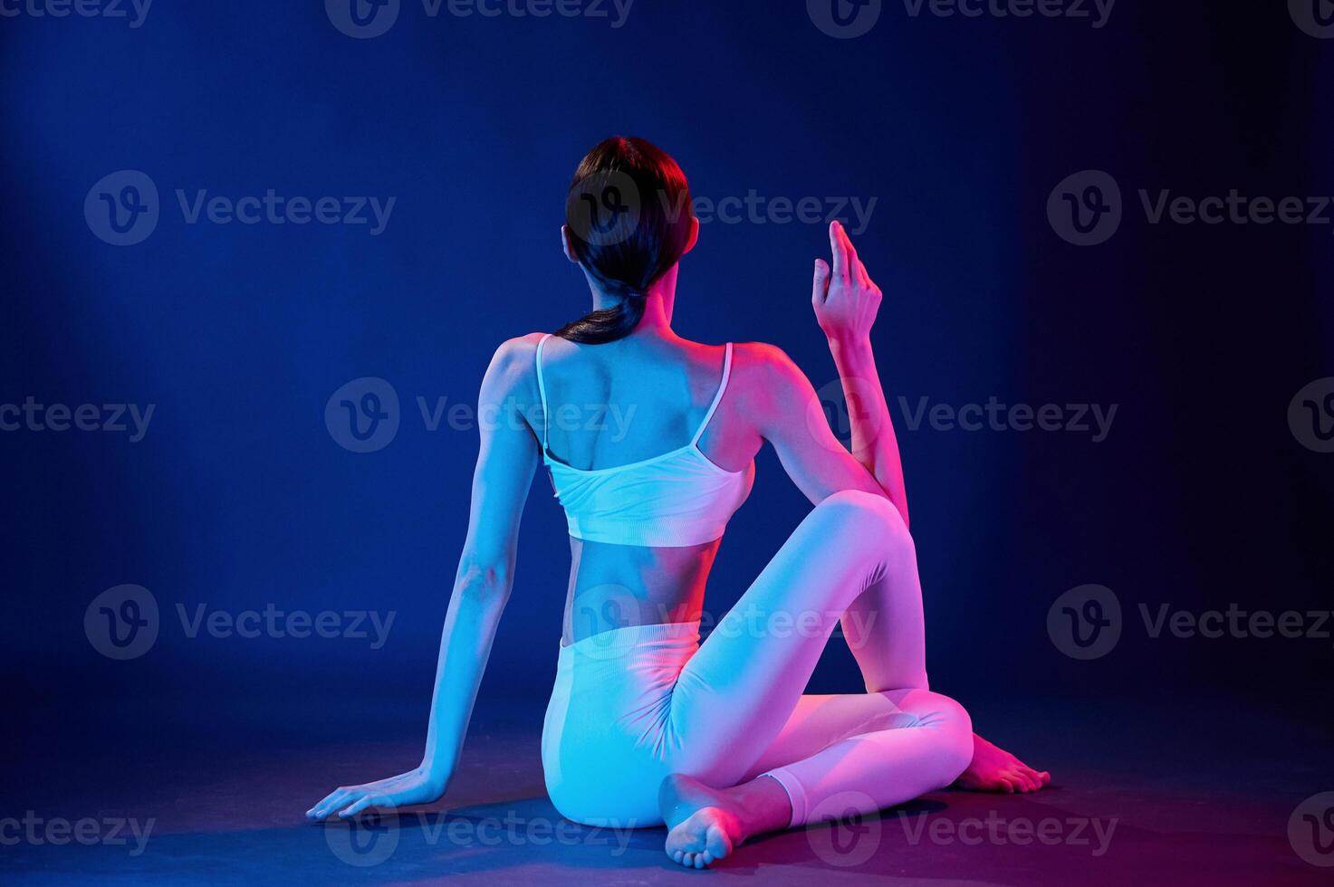 Rear view. Sitting and doing exercises. Young woman in sportive clothes is in the studio with neon lights photo