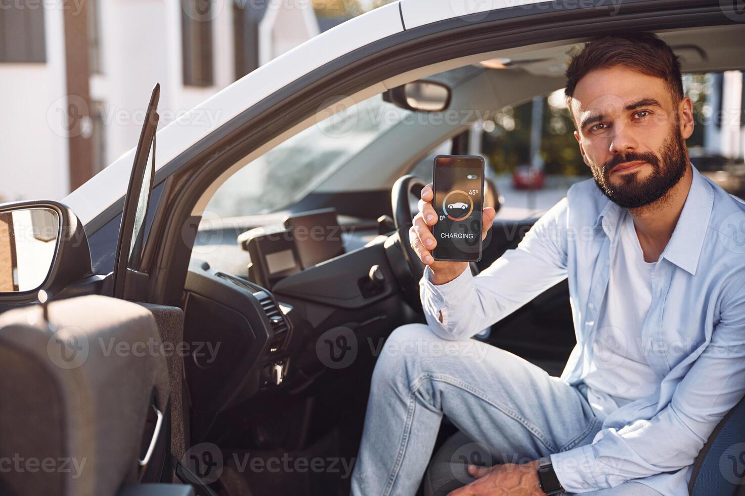 Sitting inside the vehicle, monitoring charging process by using smartphone. Young stylish man is with electric car. photo