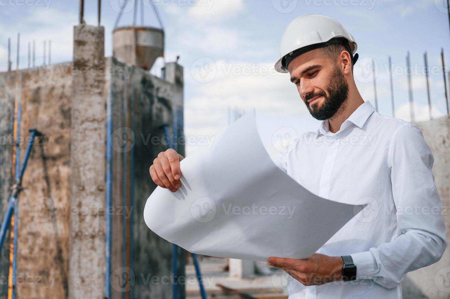Standing and holding, plan in hands. Man in uniform is working on the construction site photo