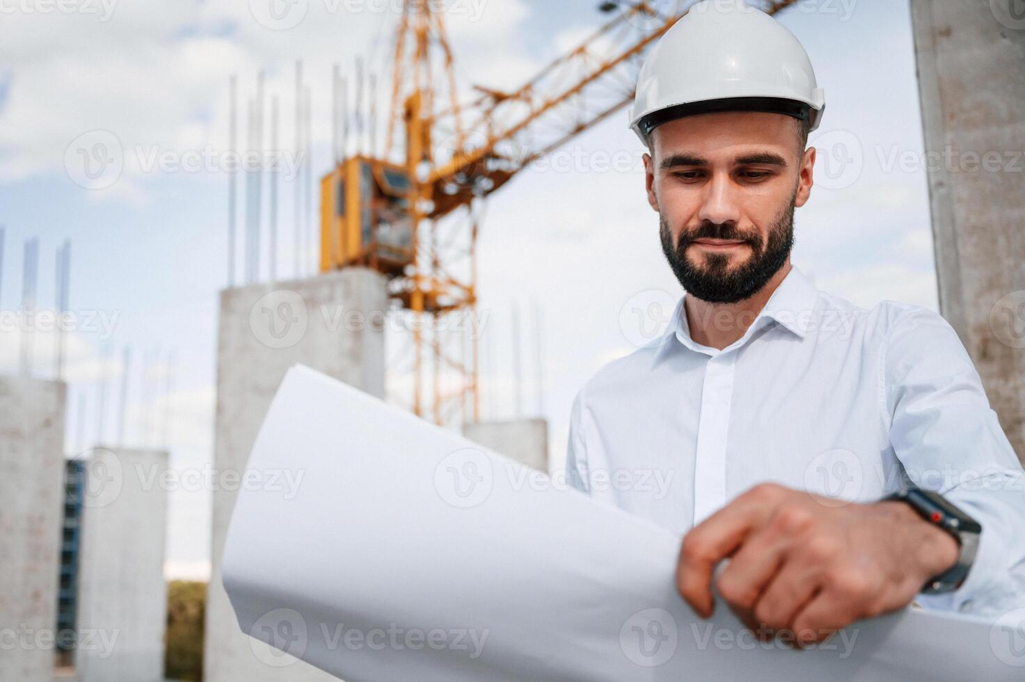 Standing and holding, plan in hands.. Man in uniform is working on the construction site photo