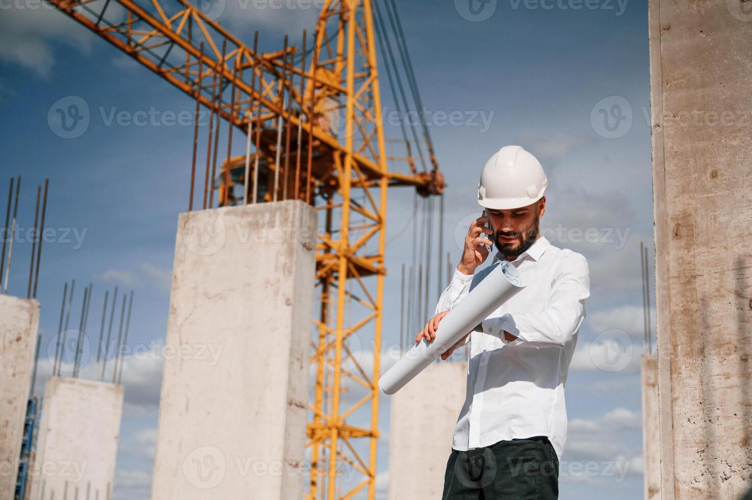 Talking by phone and holding document in hands. Man in uniform is working on the construction site photo
