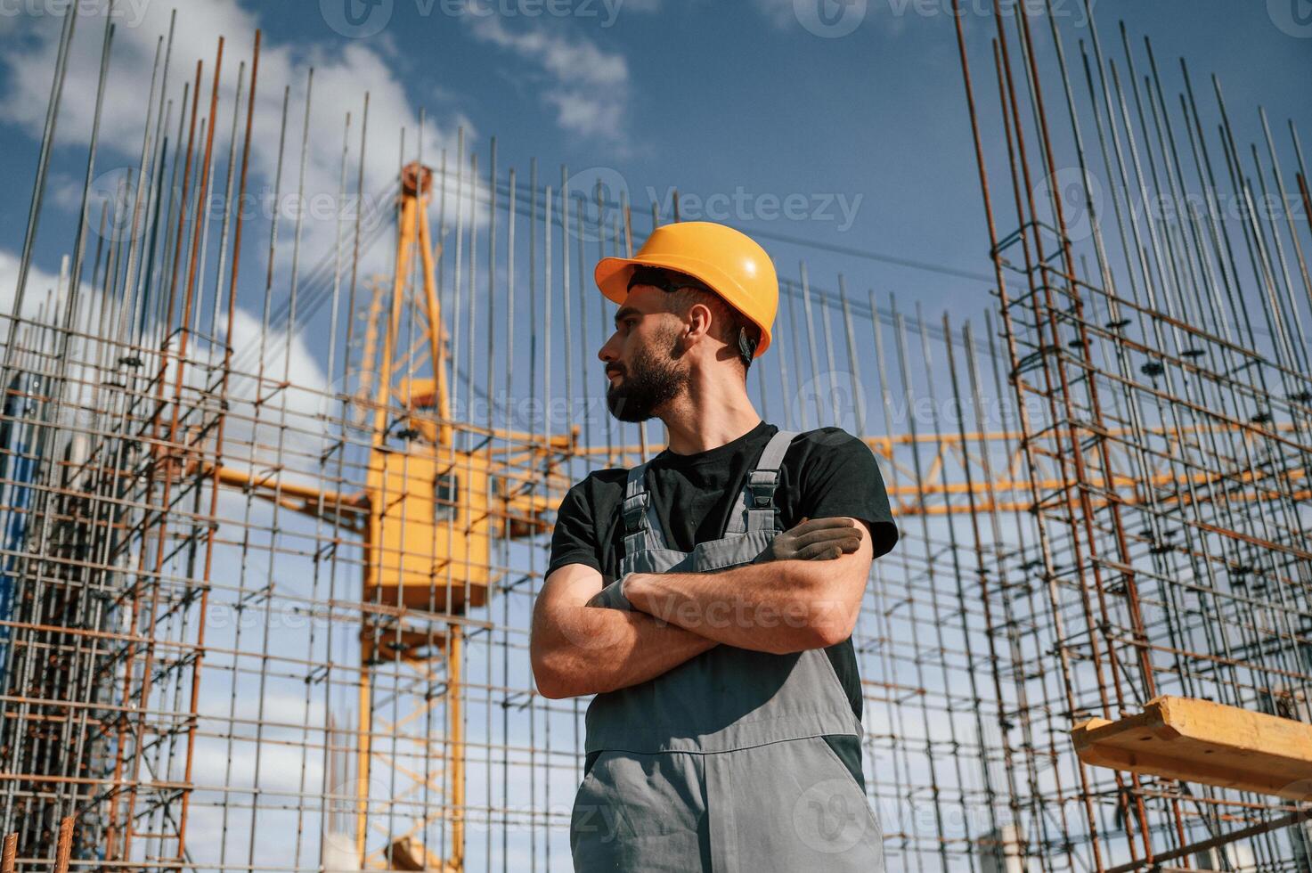 Standing against, the crane with arms crossed. Man in uniform is working on the construction site photo