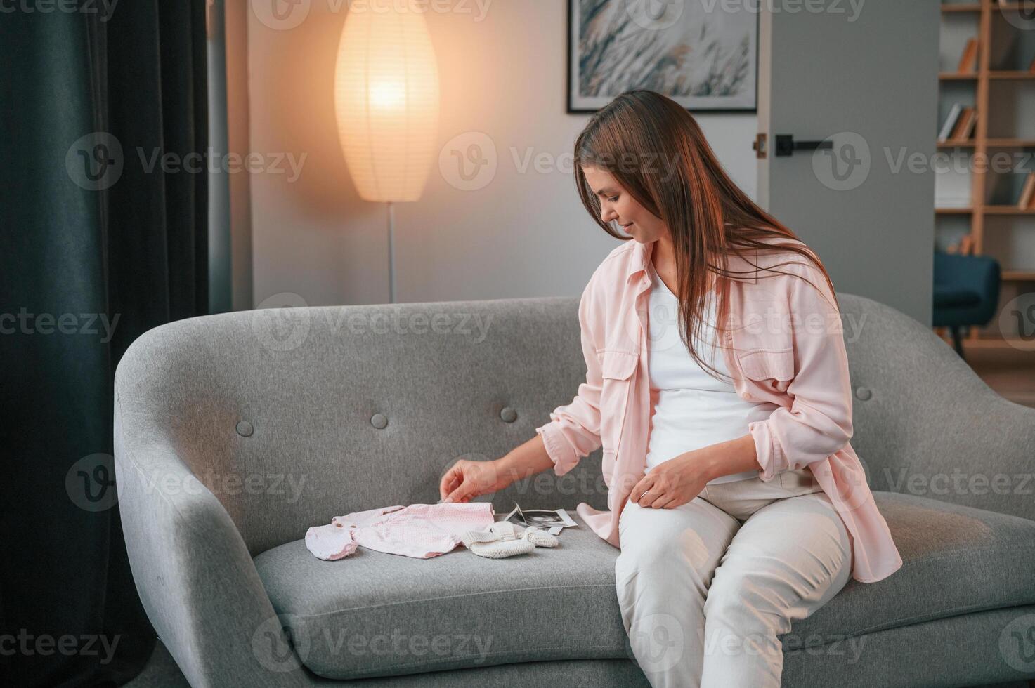 With ,little clothes for a baby on the sofa. Beautiful pregnant woman is indoors at home photo