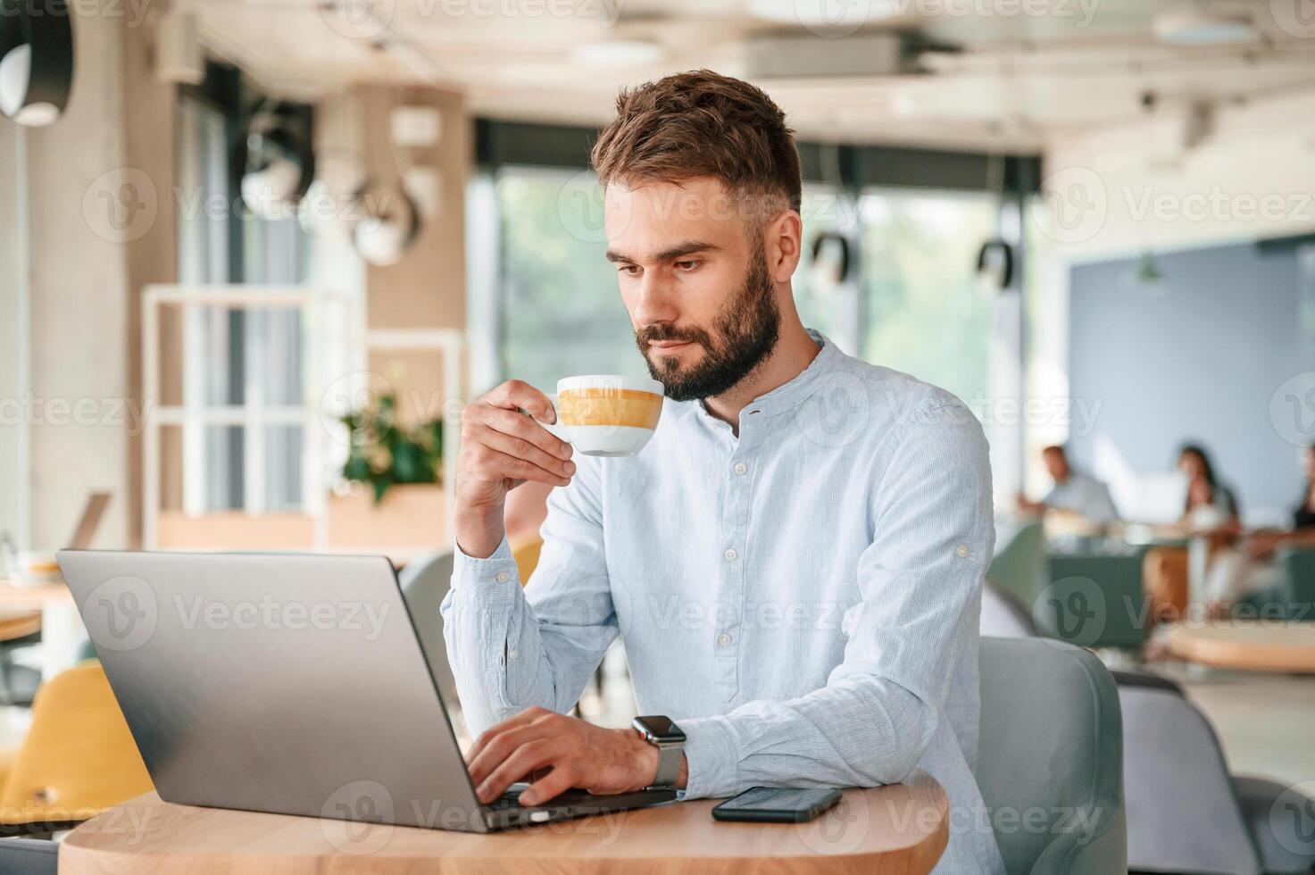 With cup of drink. Young man is sitting in the cafe and working by using laptop photo