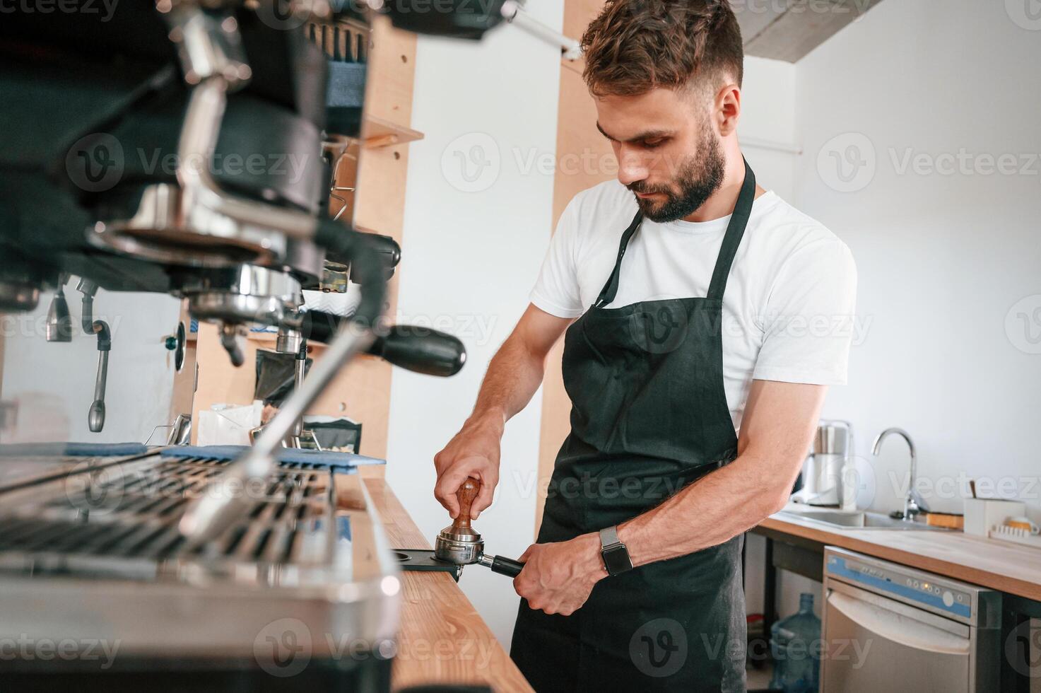 Using coffee machine.. Cafe worker in white shirt and black apron is indoors photo