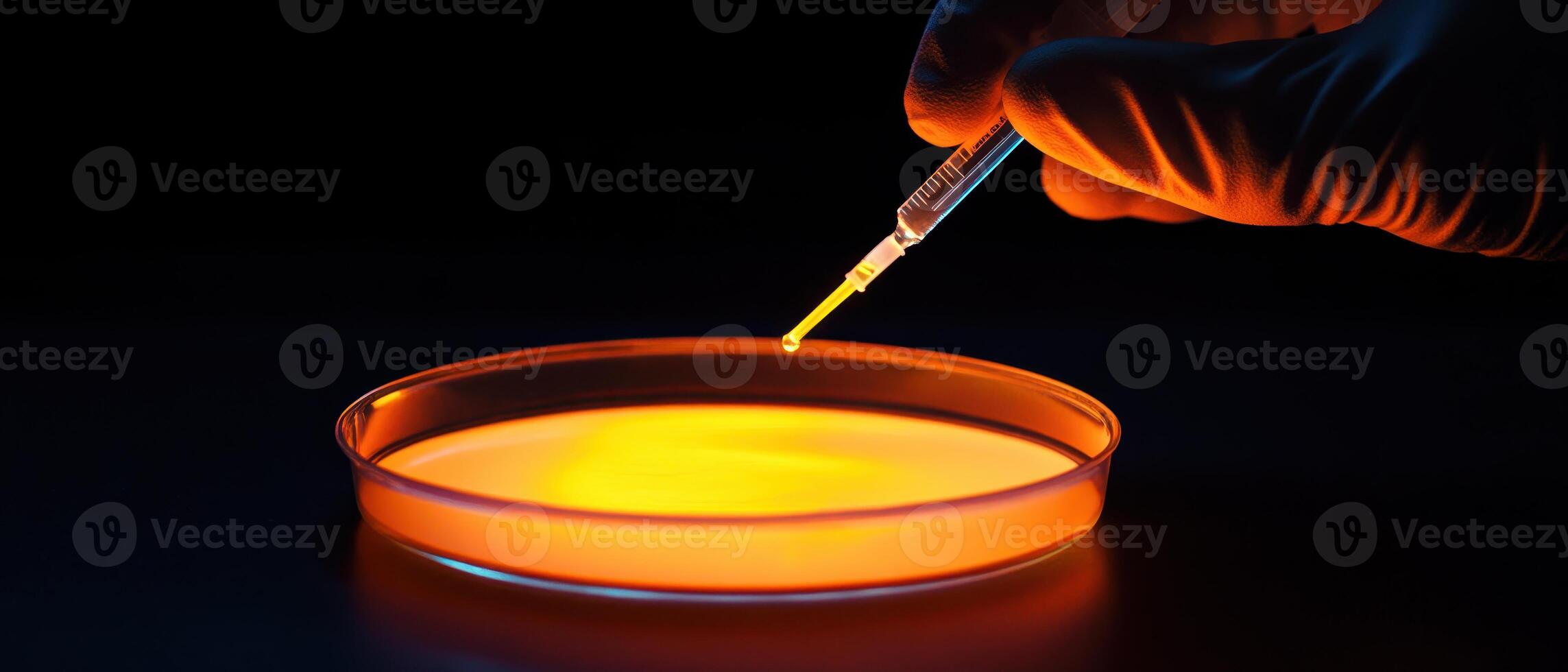 A hand in a glove holds a syringe above a glowing petri dish, suggesting a scientific experiment or laboratory setting. photo