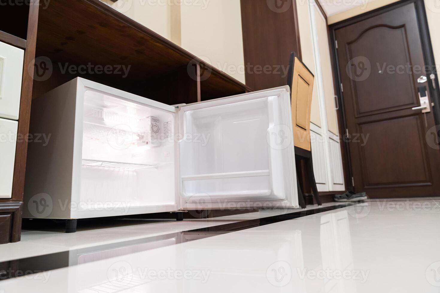 mini fridge in low angle view, inside it empty shelves placed under wooden counter and next to wood closet and chair in a luxury hotel room. photo