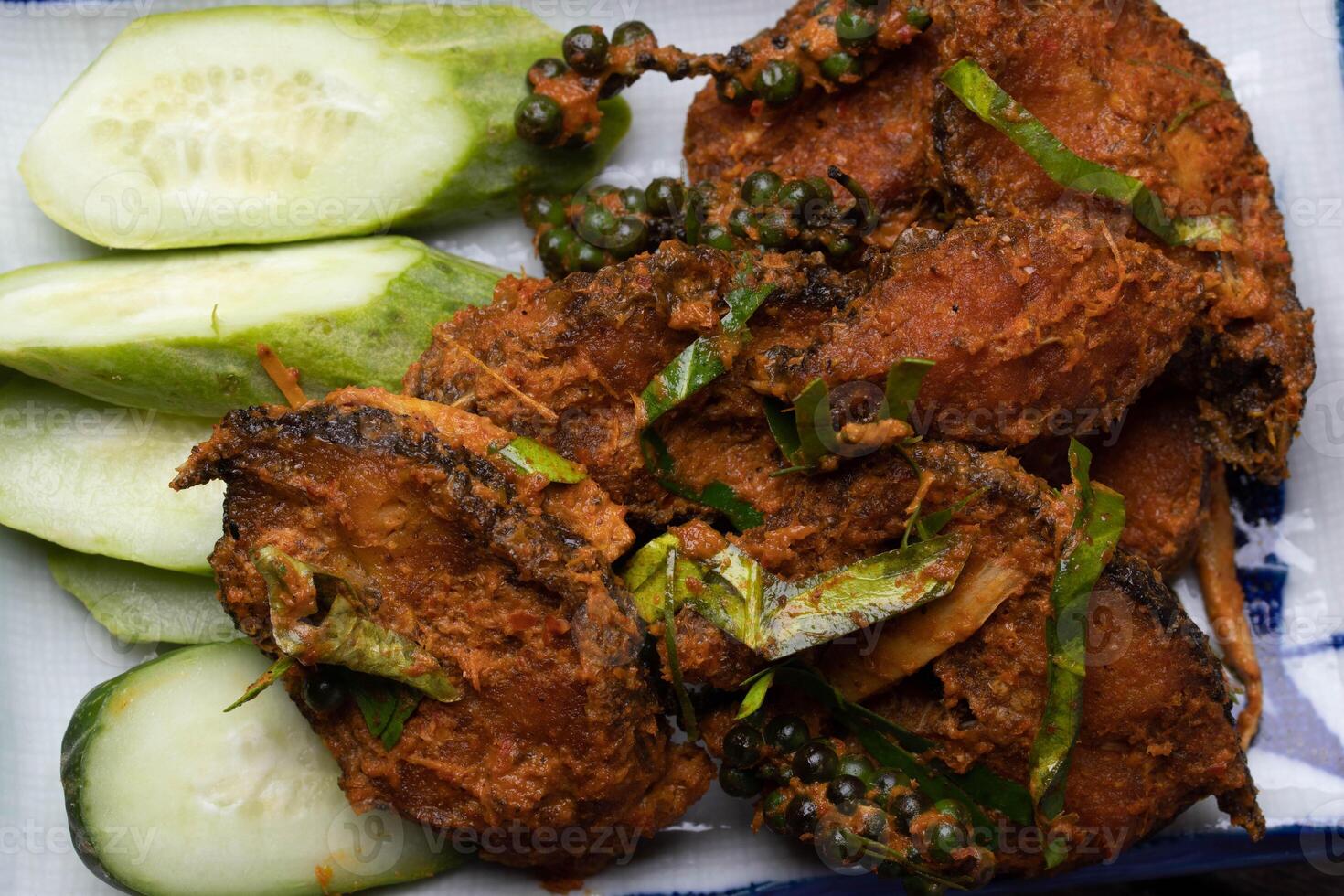 Spicy fried fish garnished with fresh herbs and served with sliced cucumbers and green peppercorns on plate. dish showcases vibrant and appetizing presentation photo