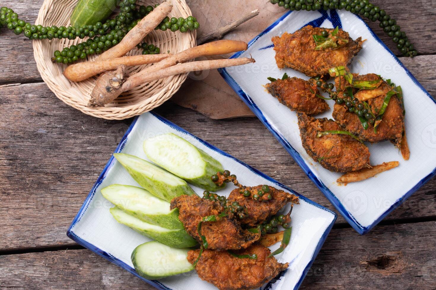 Spicy fried chicken with fresh herbs and cucumber slices on rustic wooden table. dish is garnished with green peppercorns and served alongside basket of fresh ingredients photo