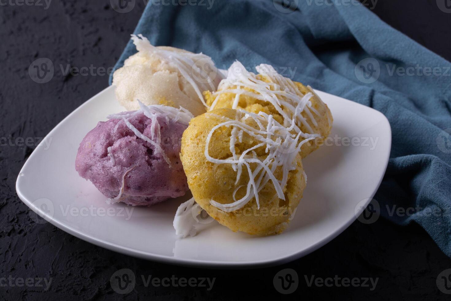 plate of colorful steamed rice cakes topped with shredded coconut, featuring purple, yellow, and white varieties, is set against dark textured background with blue cloth photo