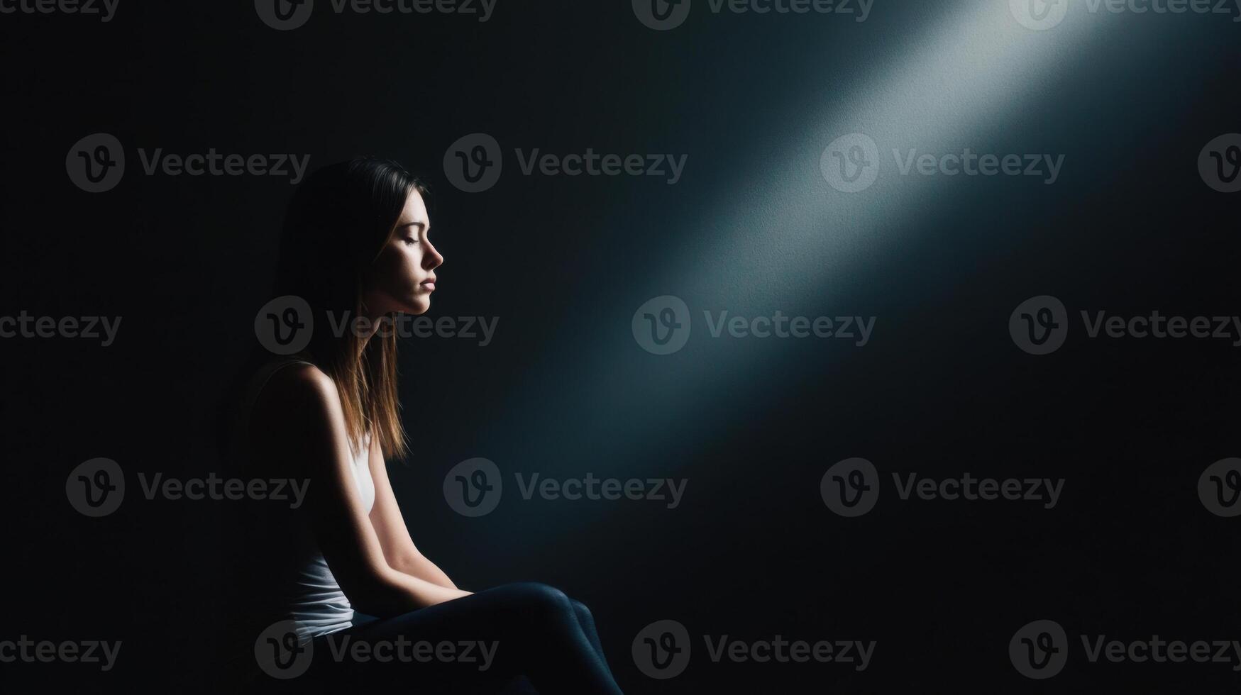 woman sits alone in dark room, illuminated by single beam of light, creating contemplative and serene atmosphere. Her expression is thoughtful and introspective photo