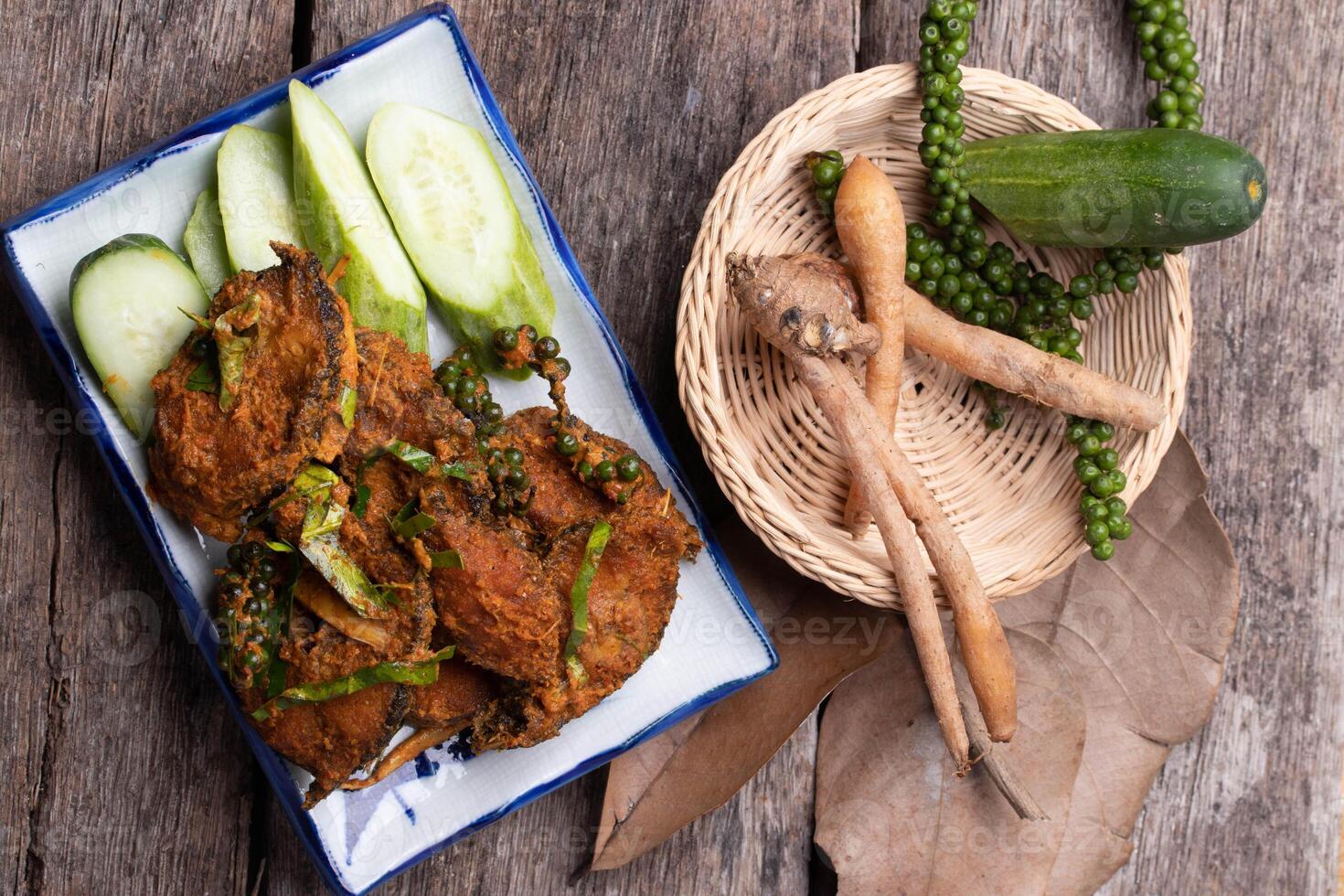 plate of spicy fried fish cakes garnished with fresh cucumber slices and green peppercorns, accompanied by basket containing fresh fingerroot and cucumber, all set on rustic wooden table photo