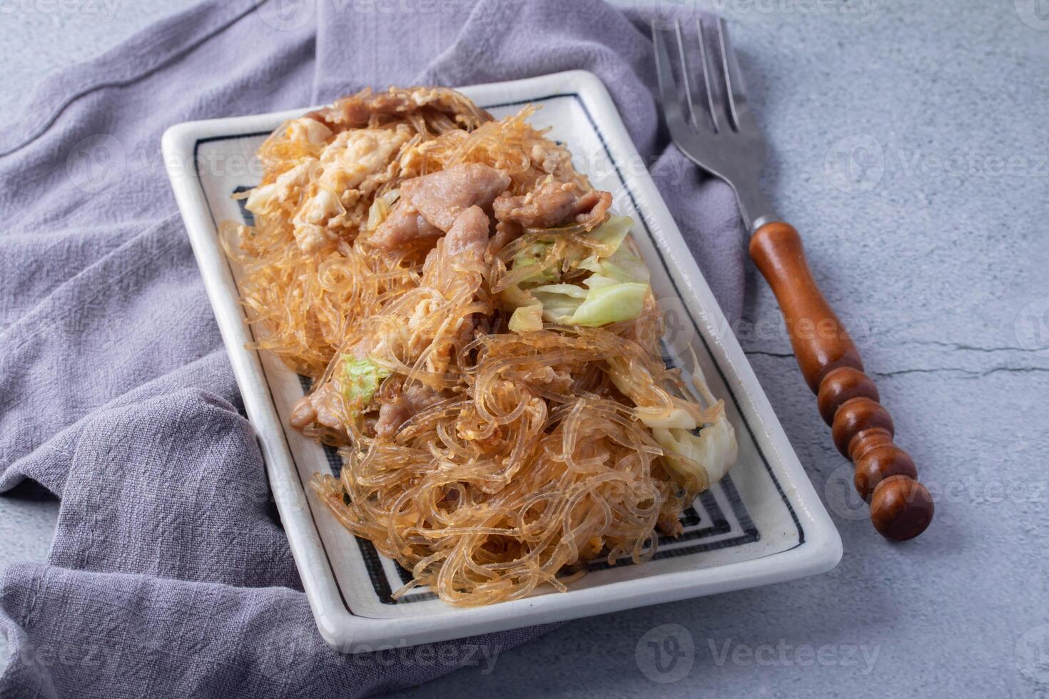 plate of stir fried glass noodles with pork and cabbage is served on rectangular dish, placed on gray cloth with wooden handled fork beside it photo