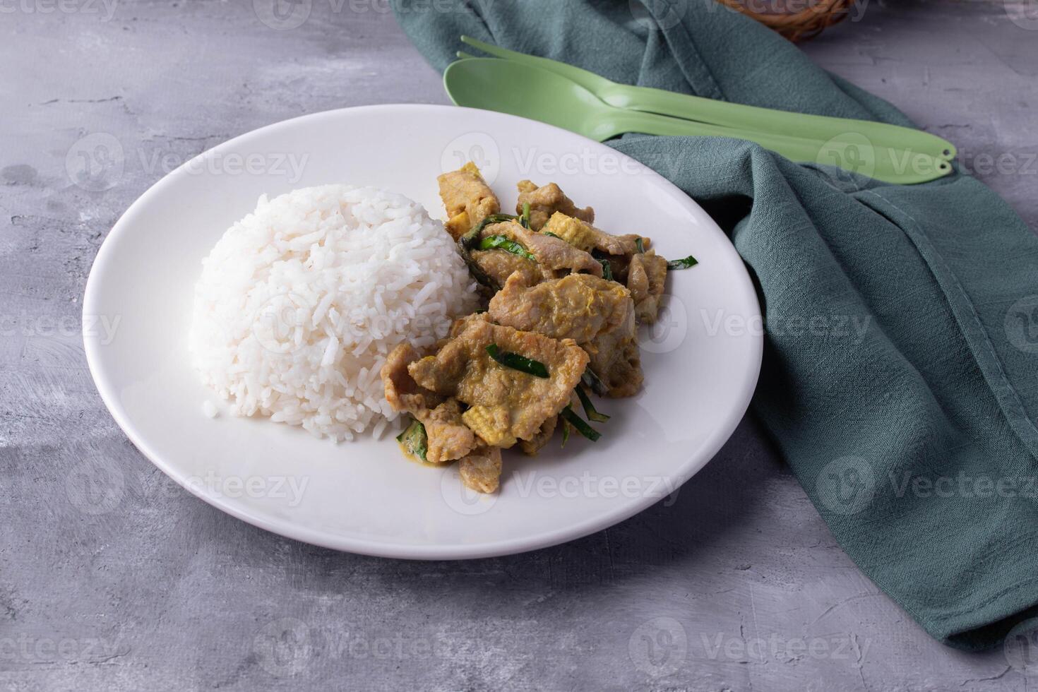 plate of white rice served with stir fried pork and vegetables, garnished with herbs, is placed on textured surface next to green cloth and utensils photo