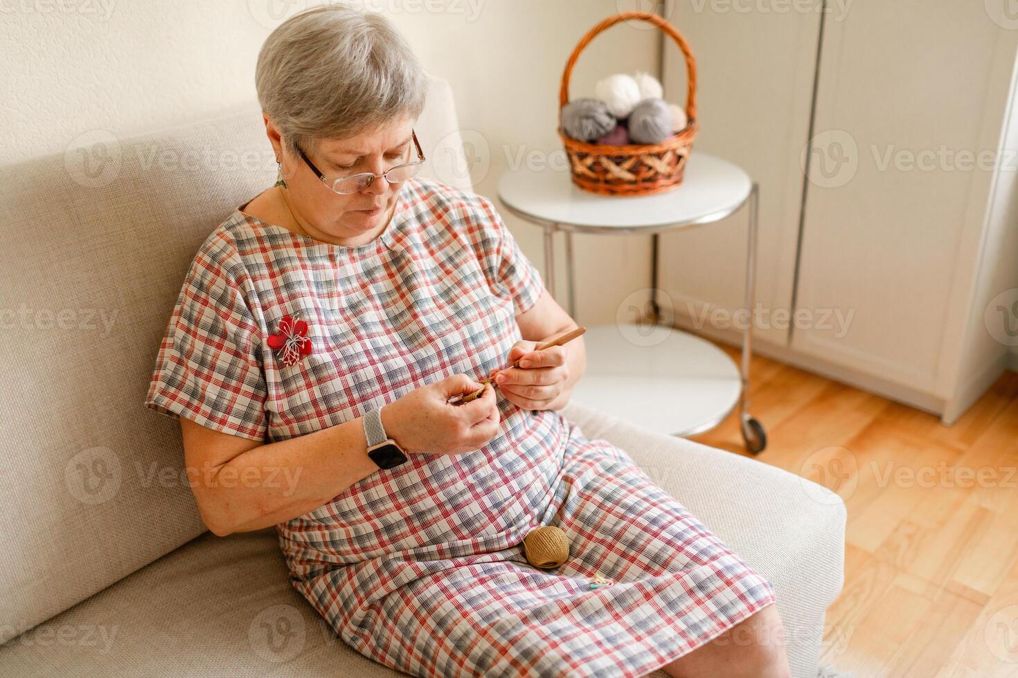 A mature woman in glasses is crocheting at home on light beige sofa photo