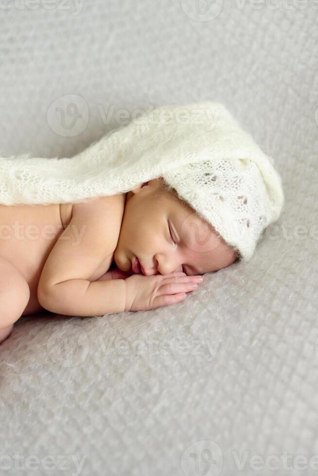 A close-up of a newborn baby girl, sleeping on a gray blanket and a white bonnet photo