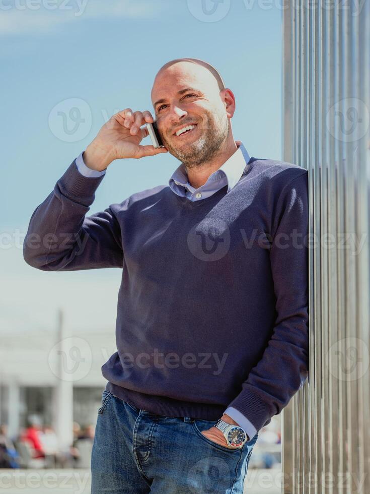 vertical photo of a businessman leaning on a wall of a building and talking to the mobile smiling