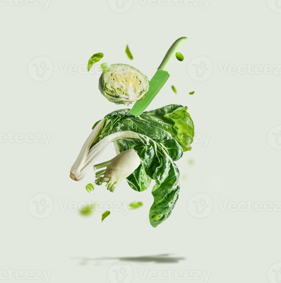 Flying green vegetables selection with knife at light background. Ingredients for healthy eating lettuce, fennel, bok choy and edamame levitating in the air. Front view. photo