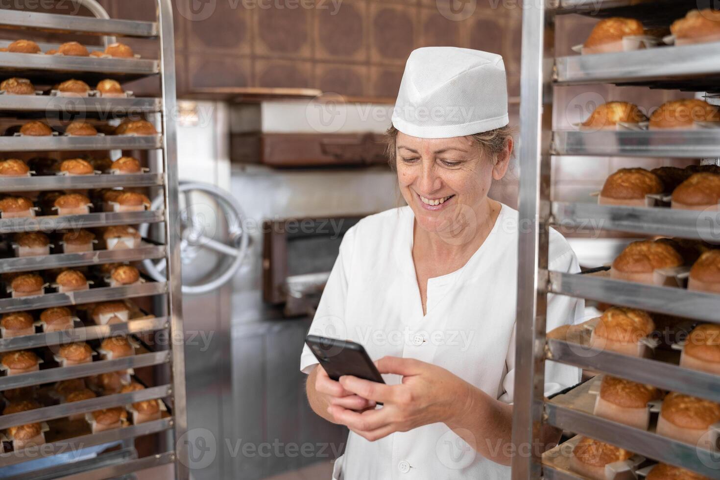 Baker woman smiling and texting with her phone surrounded by fresh baked cupcakes photo