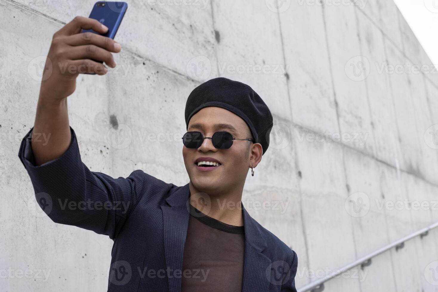 Young latin gay man with make-up on wearing a fashionable hat and sunglasses, taking selfies, isolated on a white background. LGBT. Copy space photo