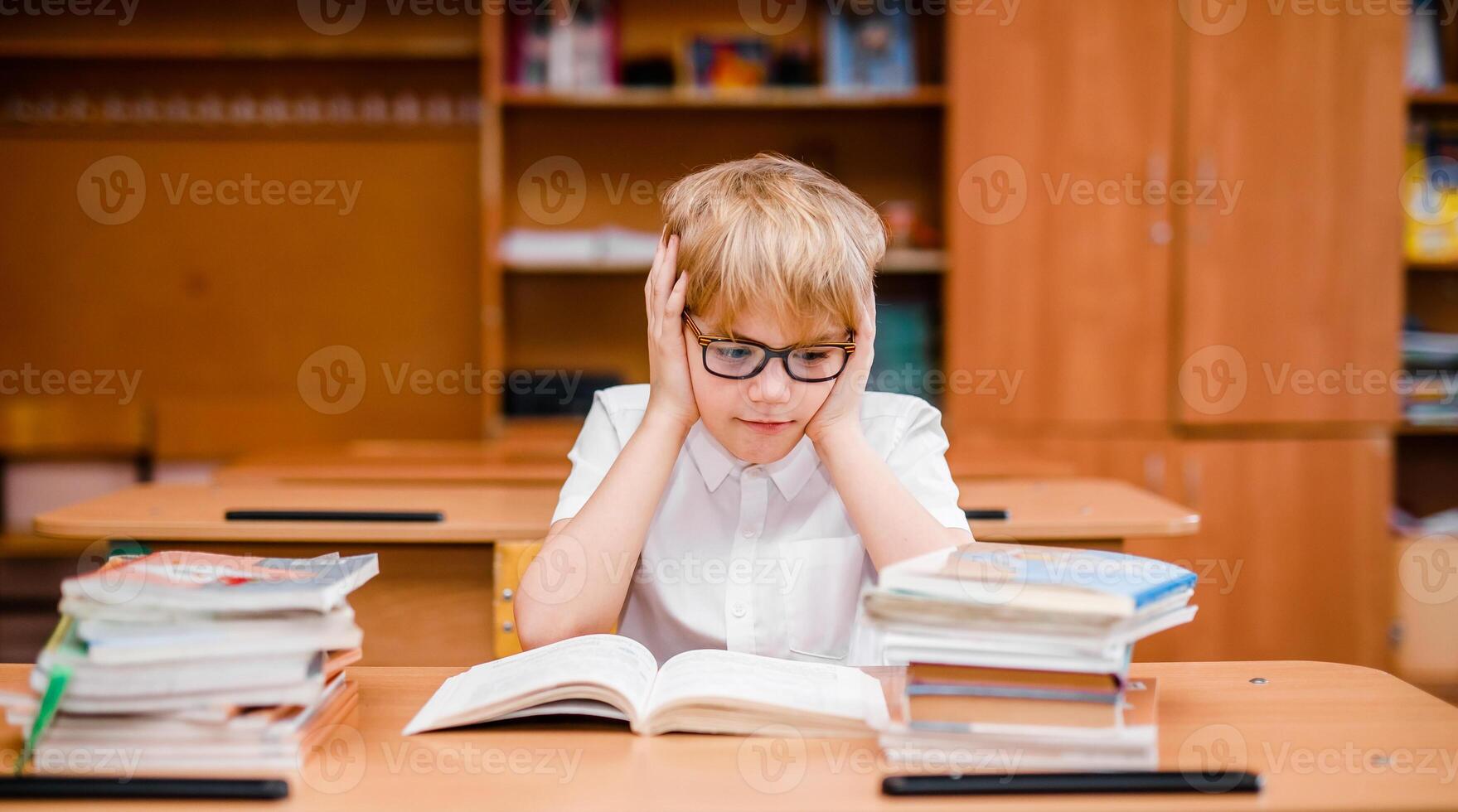 Emotional blonde boy in glasses and a white shirt at a school desk with textbooks photo