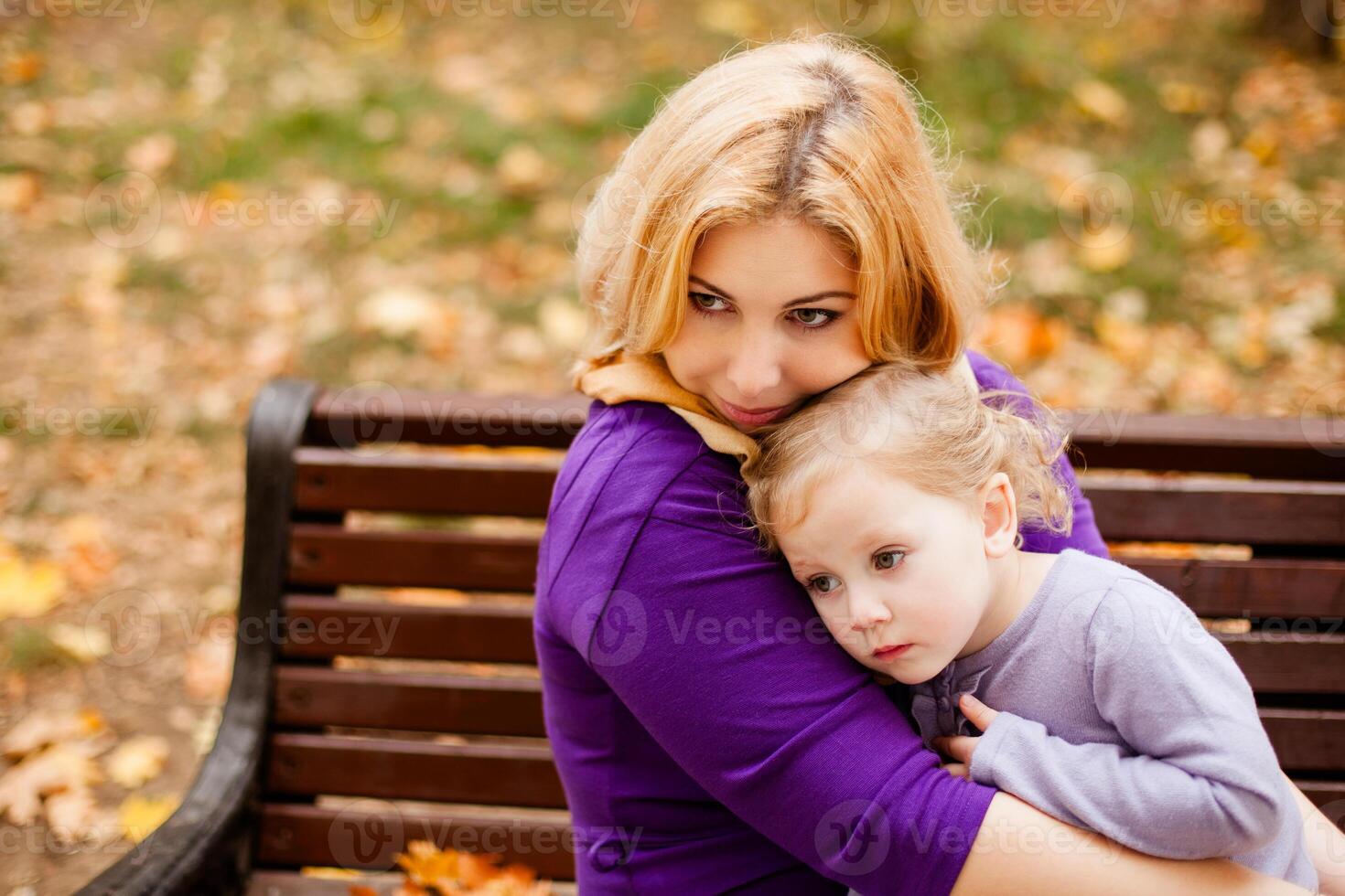 Loving mother in purple dress hugging her sad daughter on wooden park bench. photo