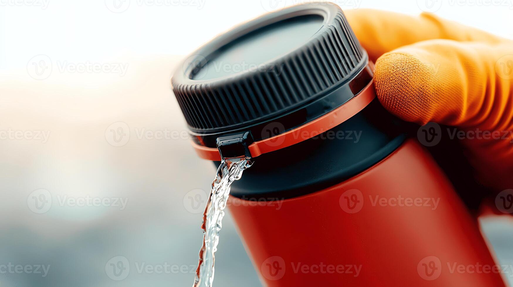 Close-up of a person in a glove pouring water from a red thermos against a blurred outdoor backdrop photo