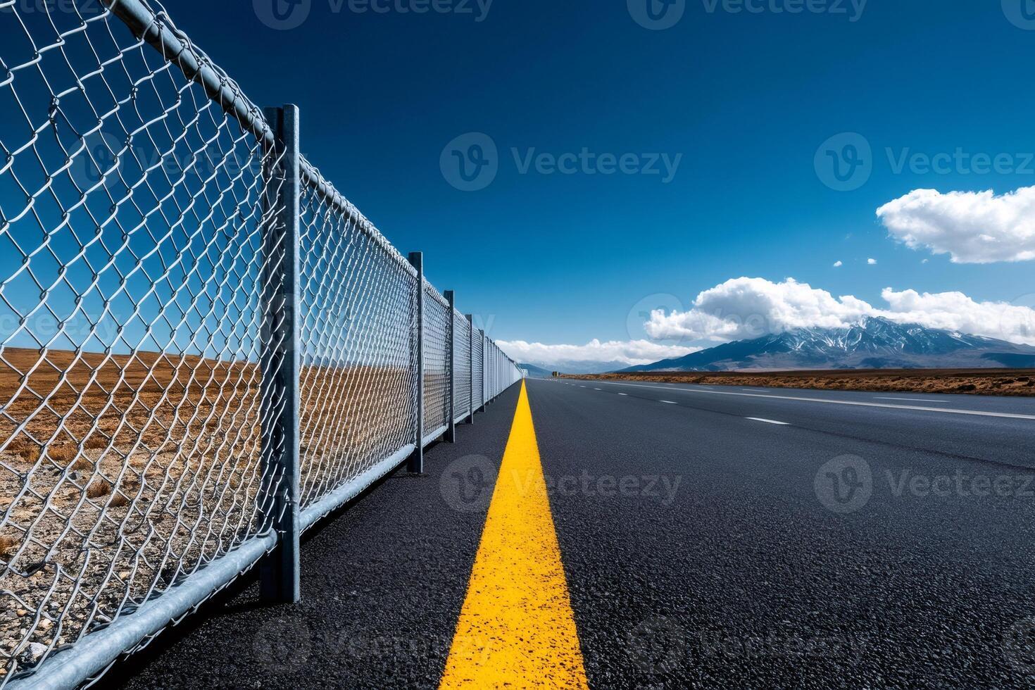 Empty asphalt road and wire mesh fence leading to mountains under blue sky photo
