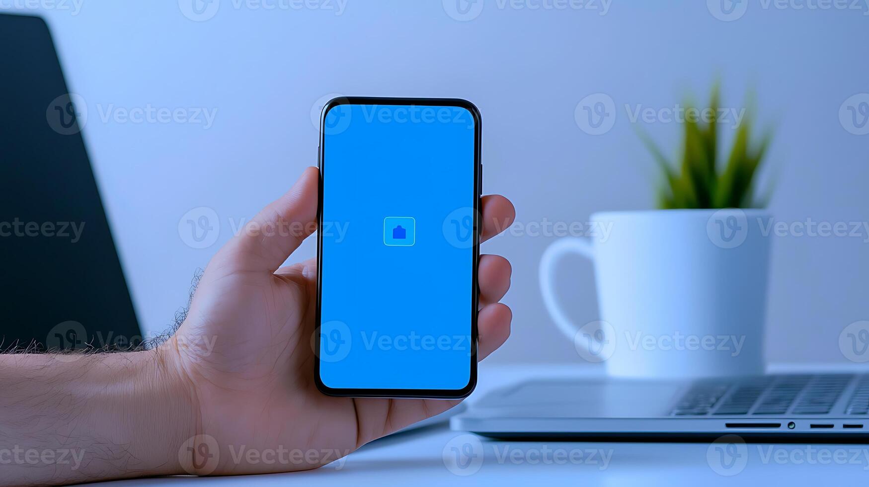 Close-up of a hand holding a smartphone with a blue screen, a laptop, and a plant in the background photo